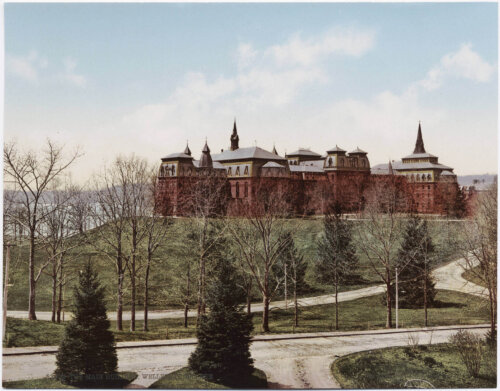 A large building on a hill with several towers with trees and a road leading up the hill.