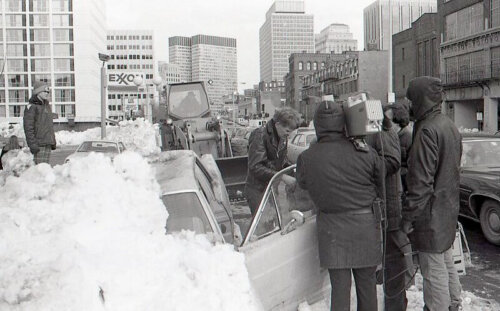 A group of men holding a tv camera standing next to a man getting out of his car which is buried in snow.