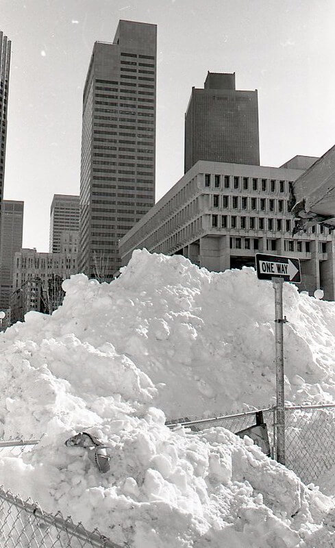 A photo of a large snow bank in front of several sky scrapers and a brutalist building in the background.