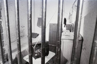 A view of a jail cell through the bars. There is a bed, tv set, and sink visible.