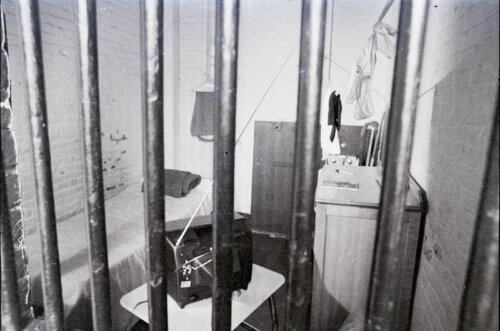 A view of a jail cell through the bars. There is a bed, tv set, and sink visible.