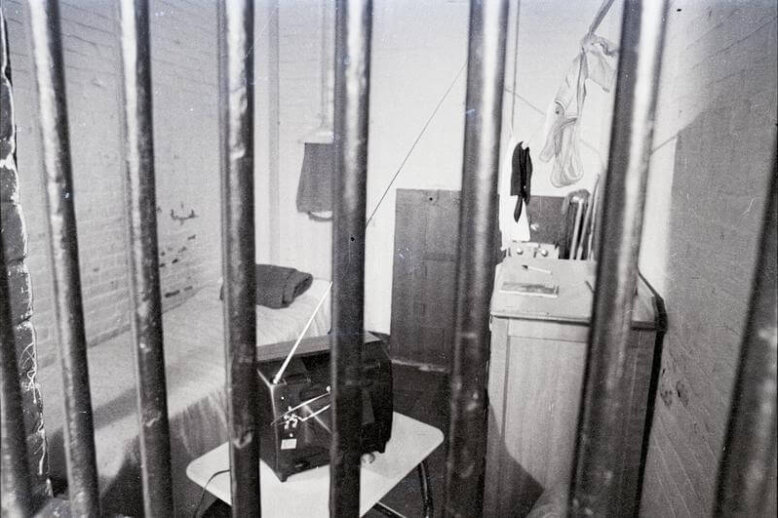 A view of a jail cell through the bars. There is a bed, tv set, and sink visible.