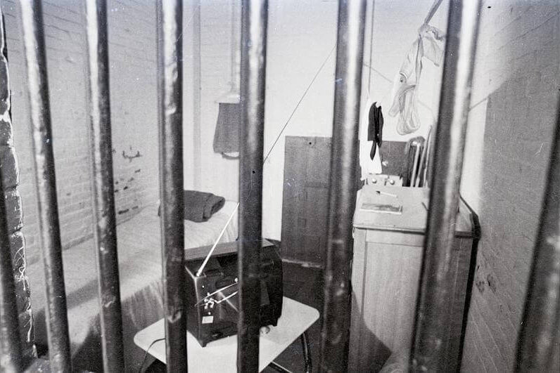 A view of a jail cell through the bars. There is a bed, tv set, and sink visible.
