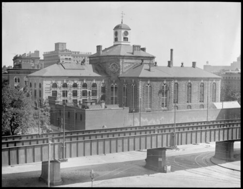 A large building with a clock tower on top with two large wings and a walled off yard. There are elevated train tracks in front of it.