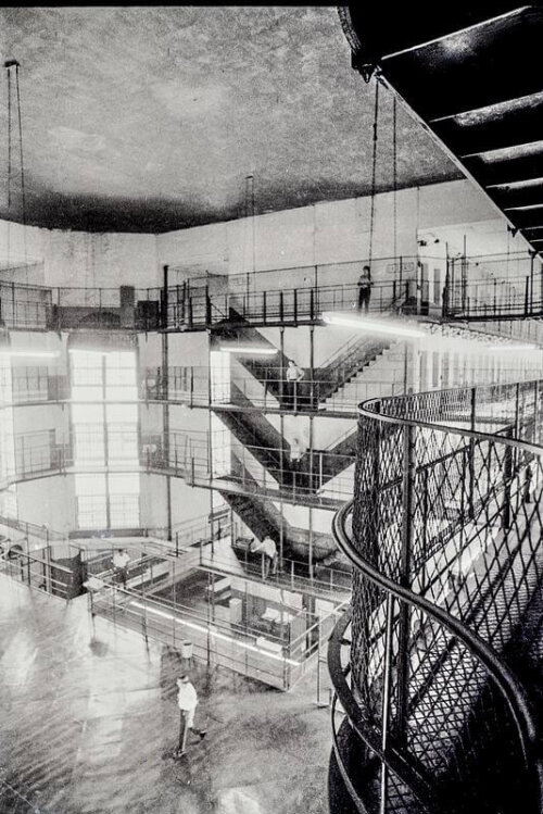 A large interior room in a jail. There are catwalks surrounding a central area which is open to the tall ceiling.