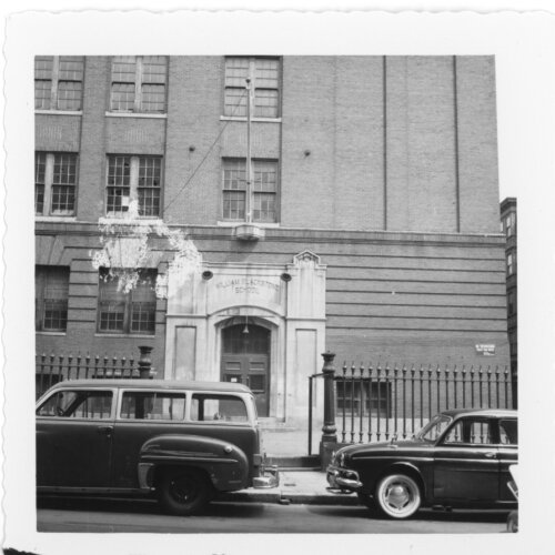 The stone entry of a school in the background. Over the door is a long flagpole. The foreground has two parked cars and a wrought iron fence.