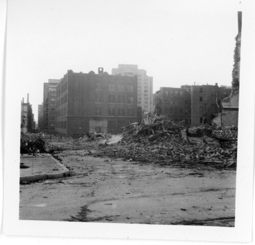 A photo of a street where several buildings have been torn down and the rubble piled in the right side of the photo. There are several larger brick buildings in the background.