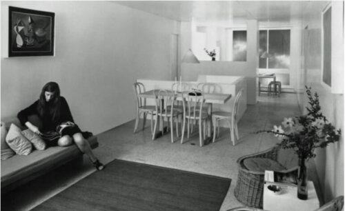 A view of an open concept apartment with a living room, dining area, and kitchen behind a half wall. A woman sits on a couch reading.