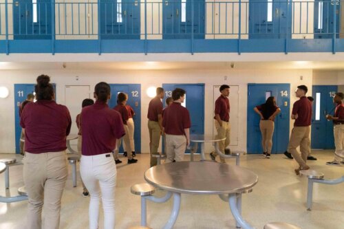 A group of teens in red shirts standing in a jail housing pod looking into cells.