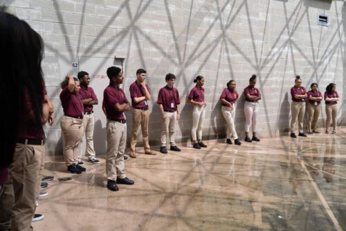 A group of teens in red shirts lean against a wall in an indoor basketball area. There are tessellated patterns from the glass roof above.