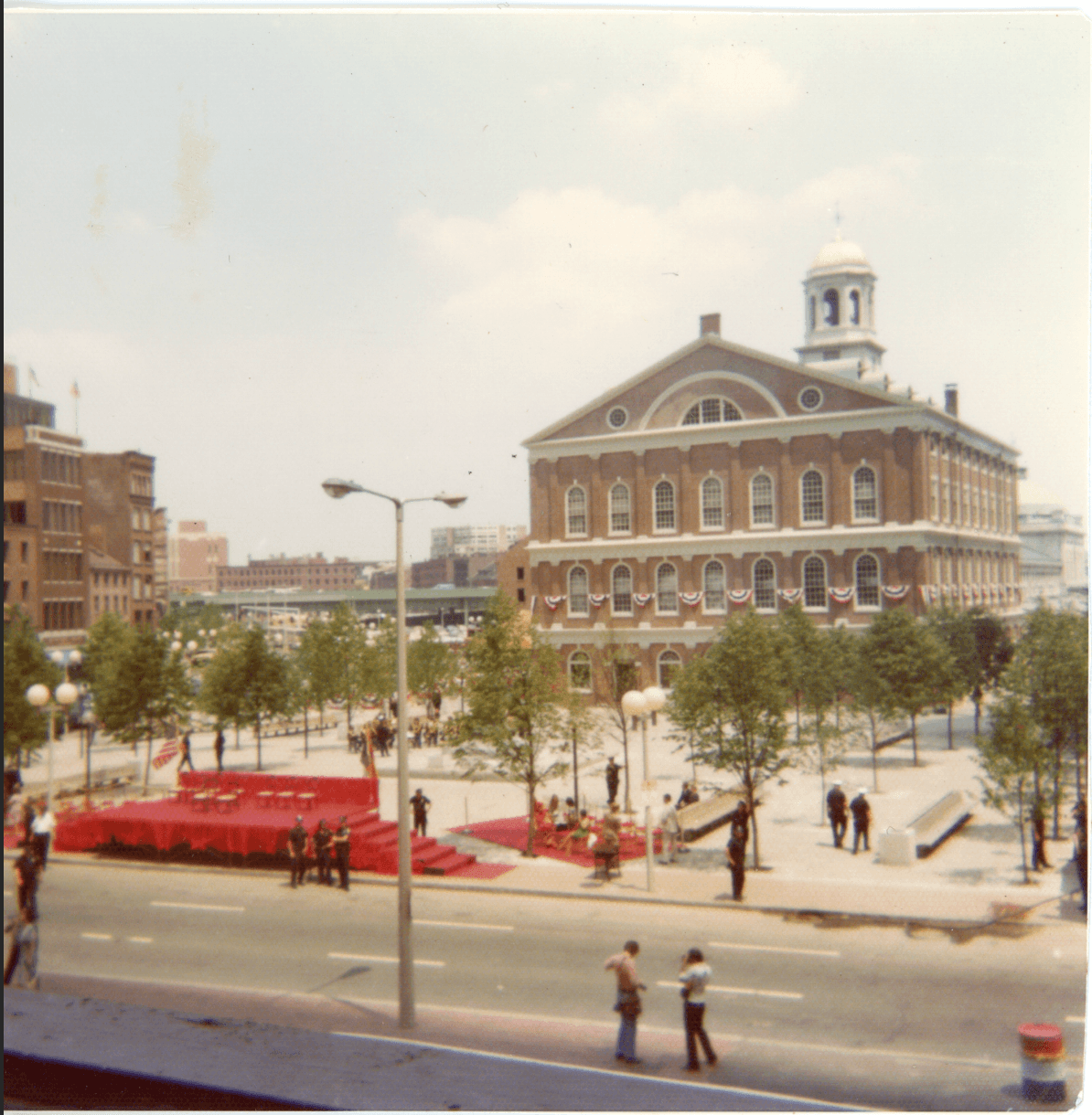 A color photograph of a four story federal building in the background with a temporary stage in a courtyard in the foreground.