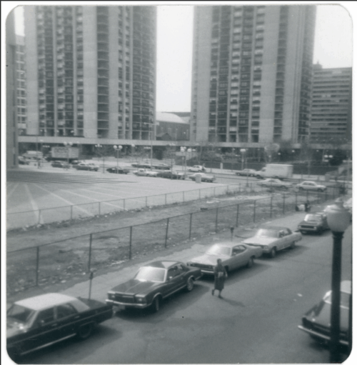A black and white photo showing a road in the lower third, a parking lot in the middle, and two large residential towers in the background.