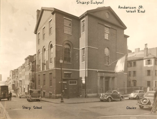 A sepia toned photo of a three story, cross-shaped school building with a large American flag over the front door. There are 1930s cars out front.