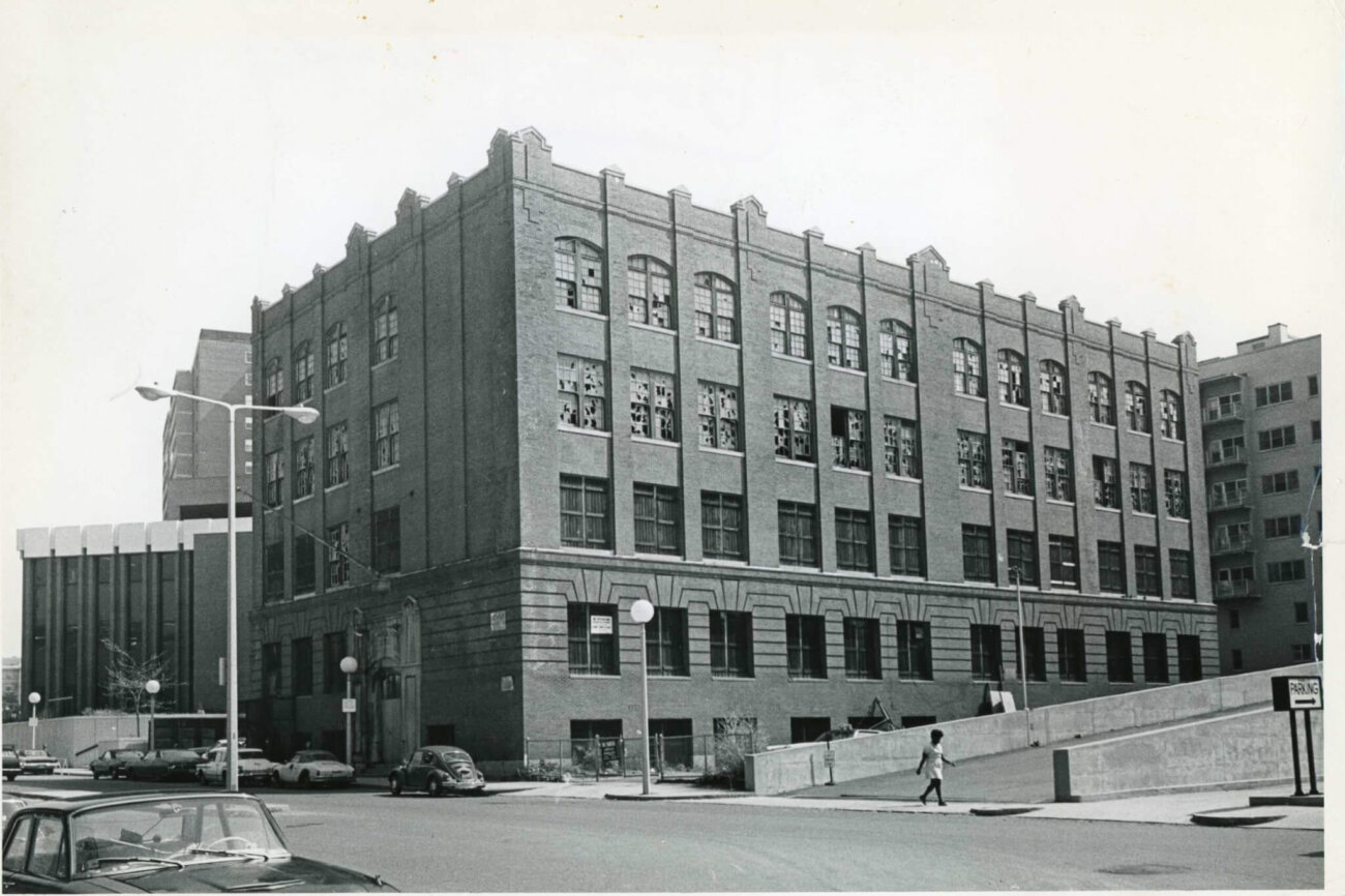 A four story brick building on a street corner with basement windows. The upper floor windows have been smashed. A woman walks along the bottom third of the image on the sidewalk.