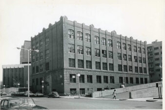 A four story brick building on a street corner with basement windows. The upper floor windows have been smashed. A woman walks along the bottom third of the image on the sidewalk.