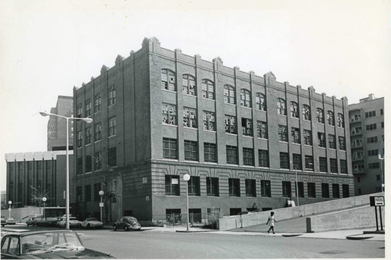 Wm_Blackstone_School_Blossom_St_1971(WEM) (1) A four story brick building on a street corner with basement windows. The upper floor windows have been smashed. A woman walks along the bottom third of the image on the sidewalk.