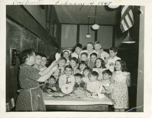 A group of children clustered around a table with crafting supplies on it while a woman in an apron and glasses shows them how to cut out a heart. They are in a classroom with a large blackboard.