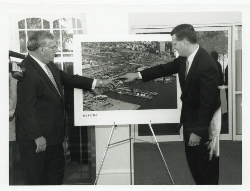 Two men in suits stand in front of a map. Both are pointing at a section of land next to a harbor.