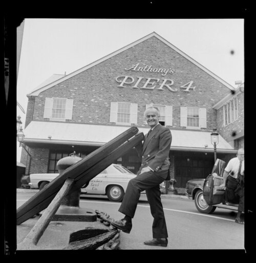 A man in a suit stands next to an anchor with a building behind him that says "Anthony's Pier 4".
