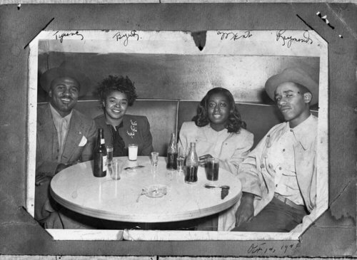 A group of four young black people sit in a corner booth drinking sodas. The photo is in a frame and signed.