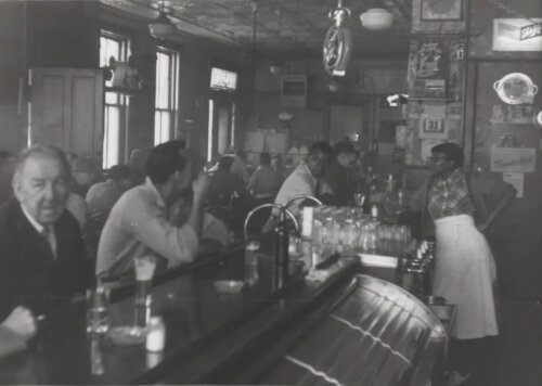 Interior of a restaurant and bar, taken from behind the bar. A man with facial hair looks at the camera while another man looks off at other customers. A waitress in an apron leans on the counter.