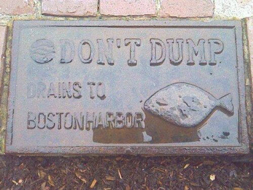 A cast iron sign on the ground that has a picture of a fish and says "Don't Dump Drains to Boston Harbor"