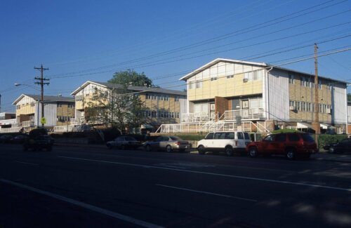 Two yellow low rise apartment buildings. They have large front porches and cars parked on the road.