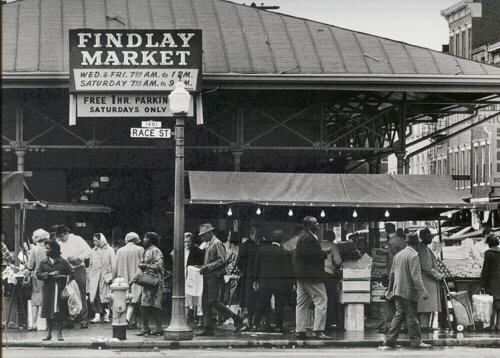 A diverse crowd at the edge of a covered public market. They carry shopping bags and crates to buy farm goods.