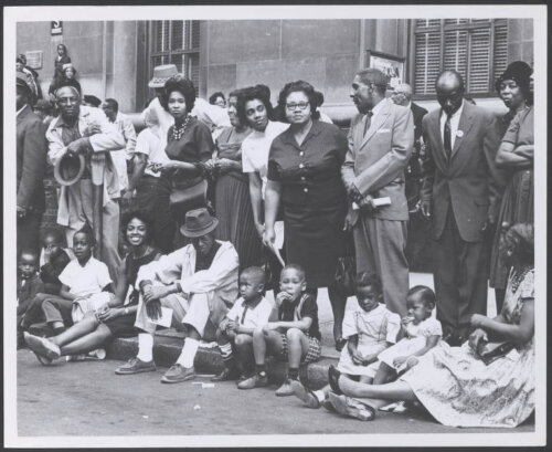 A group of black people in suits and dresses on a sidewalk. Children sit on the curb. Several people look at the camera.