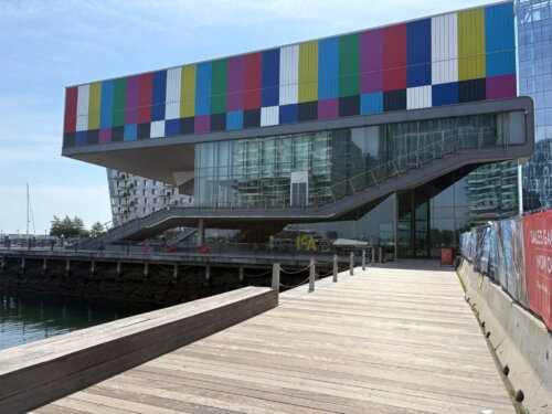 A modern building with art on the exterior to make it look like a bar code. The building is next to the water on a pier jutting out into the harbor.