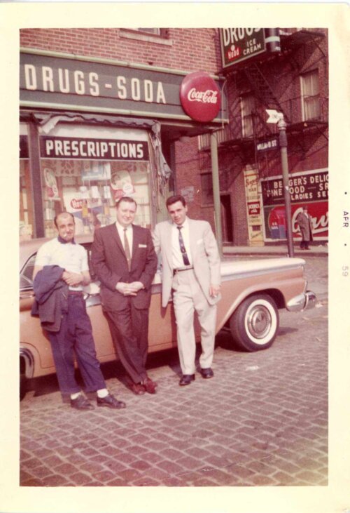 Three men leaning against a parked car in front of a drug store.