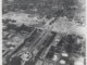 An aerial view of a neighborhood with a large train terminal at the bottom left. There is a gash through the area as buildings have been torn down for a highway construction.