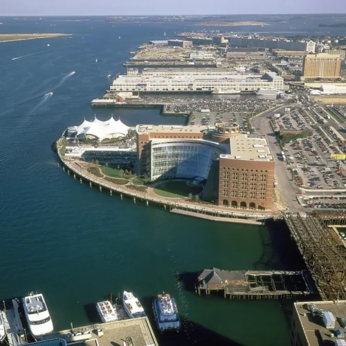 An aerial image of a U shaped building and a tent on a fan shaped piece of land. Behind the building is mostly parking lots.