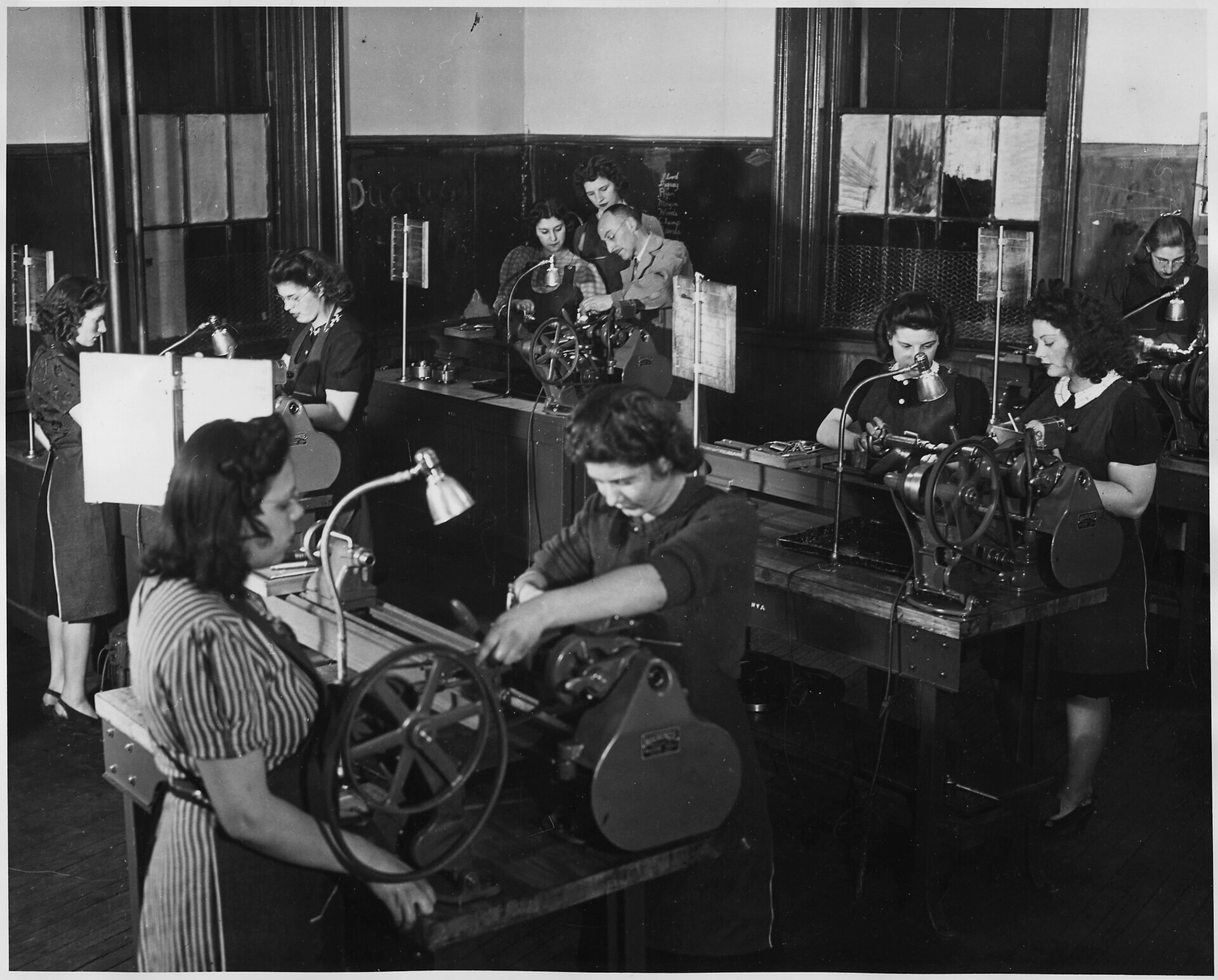 Pairs of women at machines in a factory. There is one man wearing glasses in the back corner.