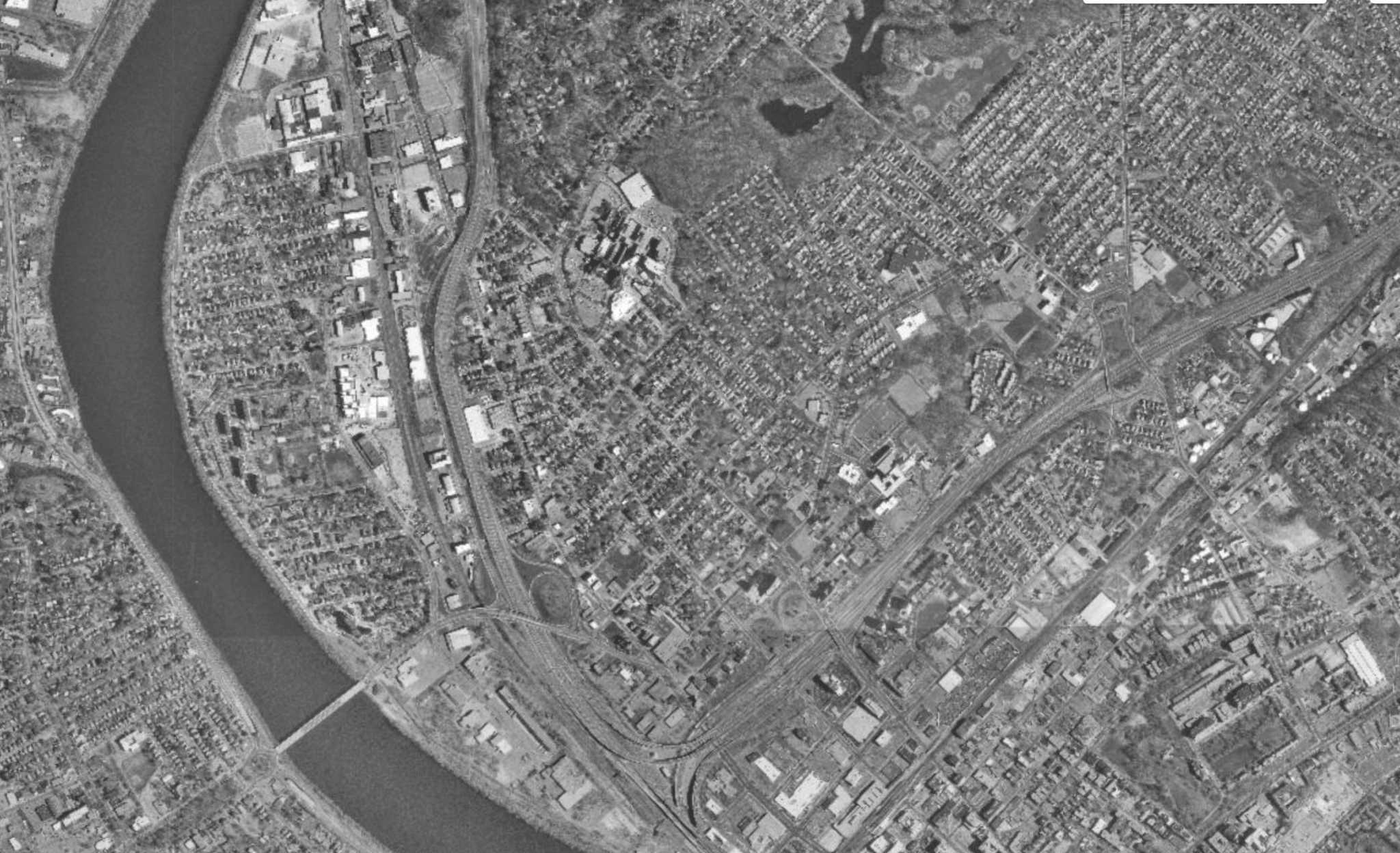 An aerial image of a city on either side of a river bend. There is a large inverted T of train tracks and a highway.