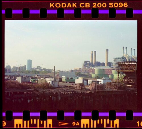 A color image visible through a photo strip that shows an industrial district with several smokestacks and detritus on the edge of a pier.