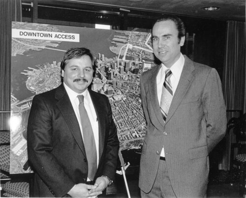 Two men in suits stand in front of a map of Downtown Boston. One man is taller than the other.