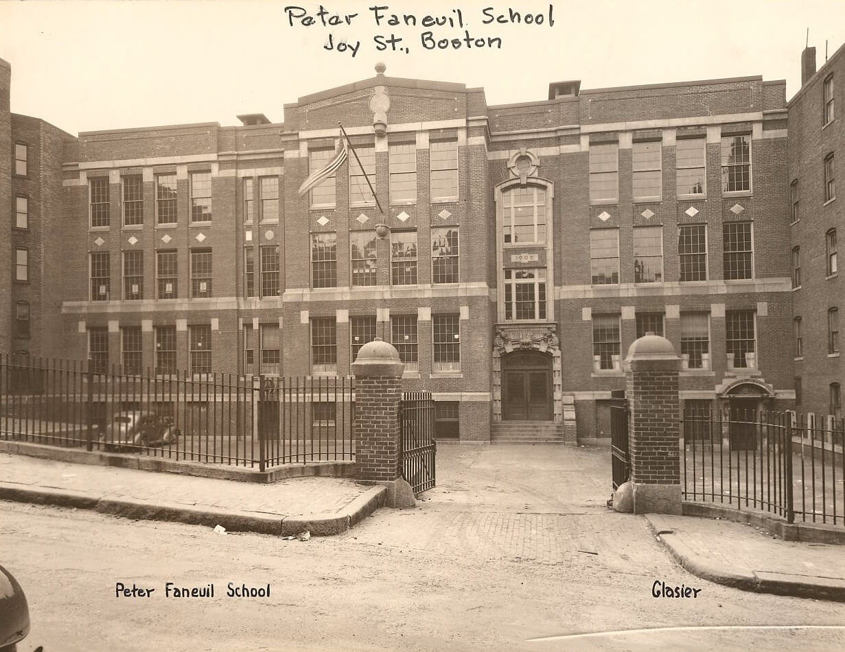 A sepia colored photo of a school behind a fence. The school is three stories and has a flag out front.