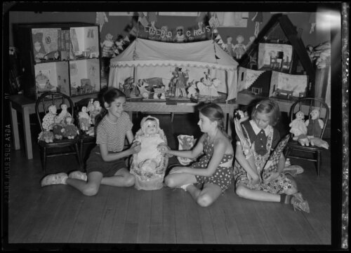 Three girls sit on the floor with several large doll houses behind them. They are playing with dolls.