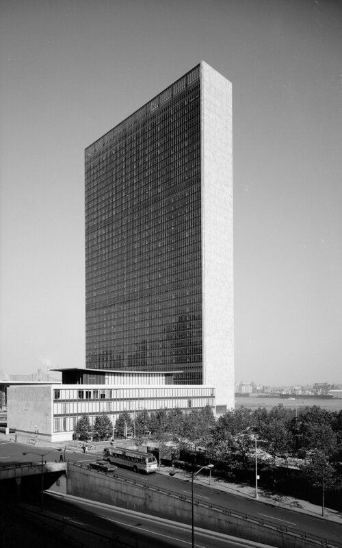 A thin tall office building with a low rise building at its base. The building over looks a river and park.