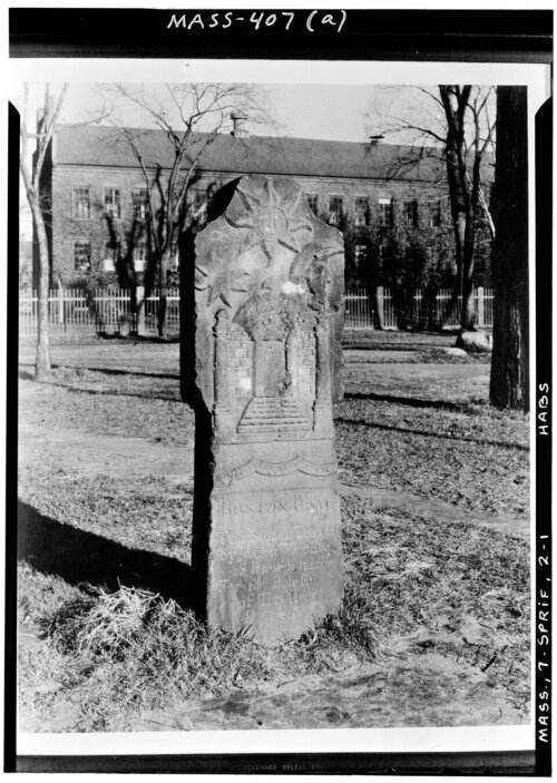 A black and white photo of a carved stone standing up in the air. The carving shows several suns.