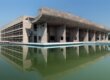 A concrete modern building with a covered deck next to a reflecting pool.