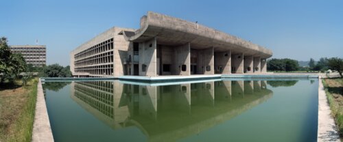 A concrete modern building with a covered deck next to a reflecting pool.