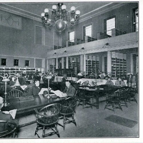 A black and white postcard showing a reading room in a library. There is a balcony above.