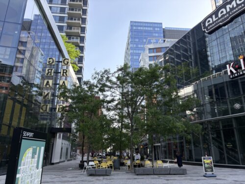 An outdoor courtyard with two tall glass buildings on either side. There are chairs and tables under small trees in the center.