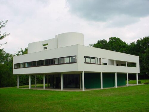 A white modern building with the outer facade supported by stilts. There is a prominent cylindrical section on the top floor.