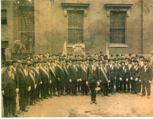 A large group of men in bowler hats and sashes standing in front of a building, some holding flags.