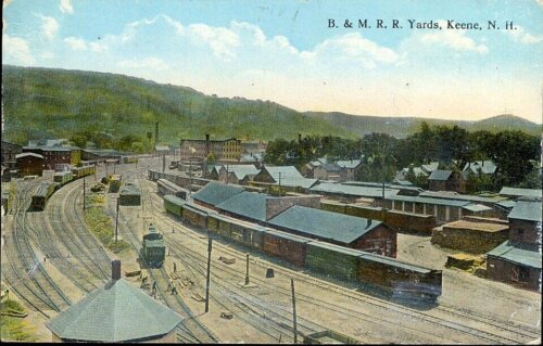 A postcard showing a train yard with several freight trains and low buildings.
