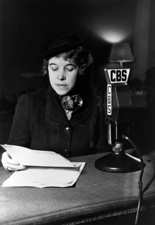 A woman reading a script sitting at a table behind a radio microphone.