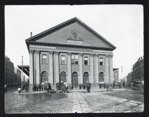 A greek revival style building with a clock in the pediment. Several carriages stand in front.